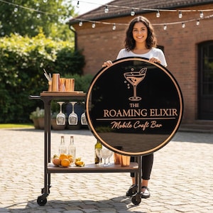 Pode incluir: Um bar artesanal m&oacute;vel com uma placa preta e cobre que diz "The Roaming Elixir Mobile Craft Bar". O carrinho de bar tem utens&iacute;lios de bar de cobre, artigos de vidro e frutas frescas. O carrinho &eacute; sobre rodas e est&aacute; ao ar livre.