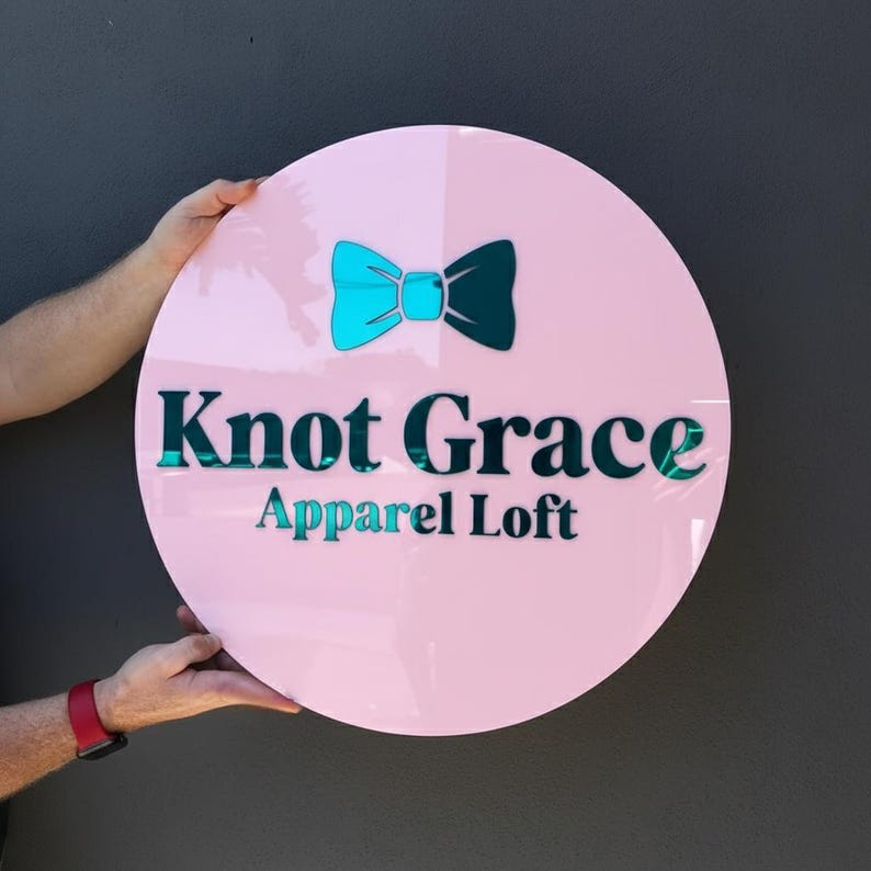 May include: A round, pink sign with the text "Knot Grace Apparel Loft" in teal. A teal and dark teal bow tie graphic is at the top. The sign is held by two hands, with a red watch visible on one wrist.