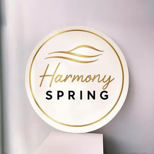 Puede incluir: Un letrero circular blanco con un borde dorado y un gráfico de onda dorada en la parte superior. El texto Harmony está escrito en letra dorada, mientras que la palabra SPRING está impresa debajo en letras mayúsculas negras y negritas.