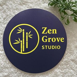 Może przedstawiać: Niebieski, okrągły znak z ż&oacute;łtymi słowami "Zen Grove STUDIO". Ż&oacute;łte koło zawiera wz&oacute;r bambusa. Znak znajduje się na białej, teksturowanej powierzchni.