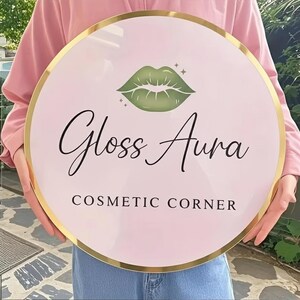 May include: A round sign with a pink background and a gold border. The sign features a green lip graphic and the words "Gloss Aura" in cursive, with "Cosmetic Corner" below in a sans-serif font.