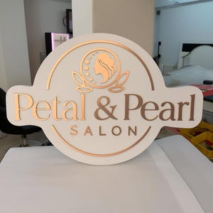 May include: A white sign with the words "Petal & Pearl SALON" in a copper color. The sign has a circular design with a woman's silhouette and floral accents. The sign is likely for a beauty salon.