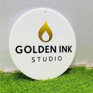 Puede incluir: Un letrero circular blanco con un icono dorado en forma de llama en la parte superior, con el texto negro GOLDEN INK STUDIO impreso debajo. El letrero tiene un pequeño orificio en el centro superior para colgar y una superficie lisa.