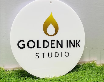 以下が含まれることがあります： 上部に金色の炎の形をしたアイコン、その下に黒い文字で「GOLDEN INK STUDIO」と印刷された円形の白い看板。看板の上部中央には吊り下げ用の小さな穴があり、表面は滑らかで平らな仕上がりになっています。