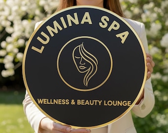 May include: A round black sign with gold lettering and trim. The sign reads "LUMINA SPA" at the top and "WELLNESS & BEAUTY LOUNGE" at the bottom. A gold line drawing of a woman's face is in the center.