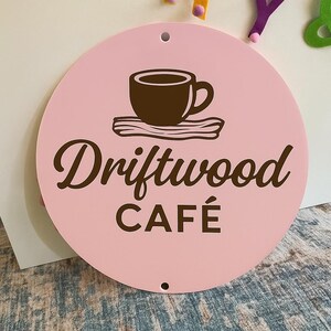 Może przedstawiać: Okrągły, r&oacute;żowy znak z napisem "Driftwood CAF&Eacute;" w brązowym skrypcie. Brązowa grafika filiżanki do kawy znajduje się nad tekstem. Znak ma dwa małe otwory u g&oacute;ry i u dołu.