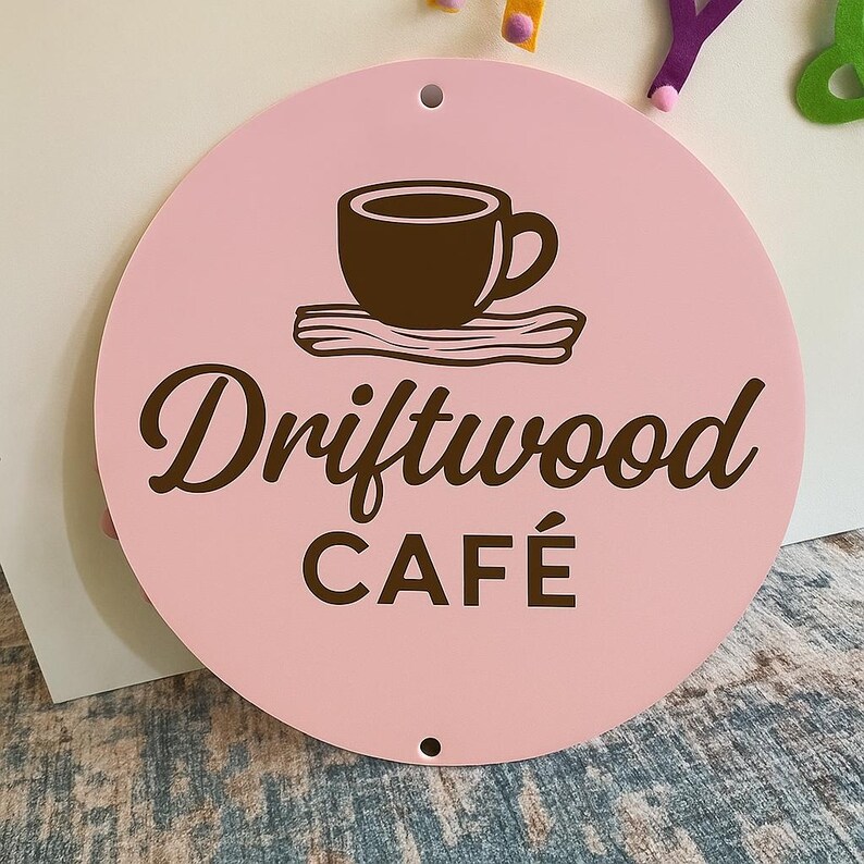 Może przedstawiać: Okrągły, r&oacute;żowy znak z napisem "Driftwood CAF&Eacute;" w brązowym skrypcie. Brązowa grafika filiżanki do kawy znajduje się nad tekstem. Znak ma dwa małe otwory u g&oacute;ry i u dołu.