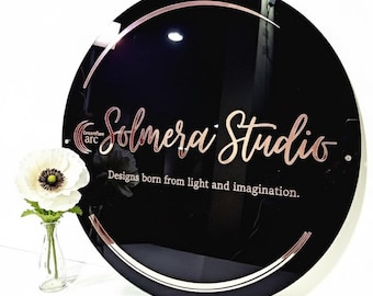 May include: A round black sign with rose gold lettering and trim. The sign reads "Solmera Studio" with the tagline "Designs born from light and imagination." A small vase with a white flower is in the lower left corner.