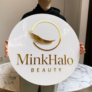 Może przedstawiać: Okrągły biały znak ze złotymi literami i złotym wzorem rzęs. Znak głosi "MinkHalo BEAUTY" w eleganckiej czcionce. Wz&oacute;r rzęs znajduje się w złotym okręgu.