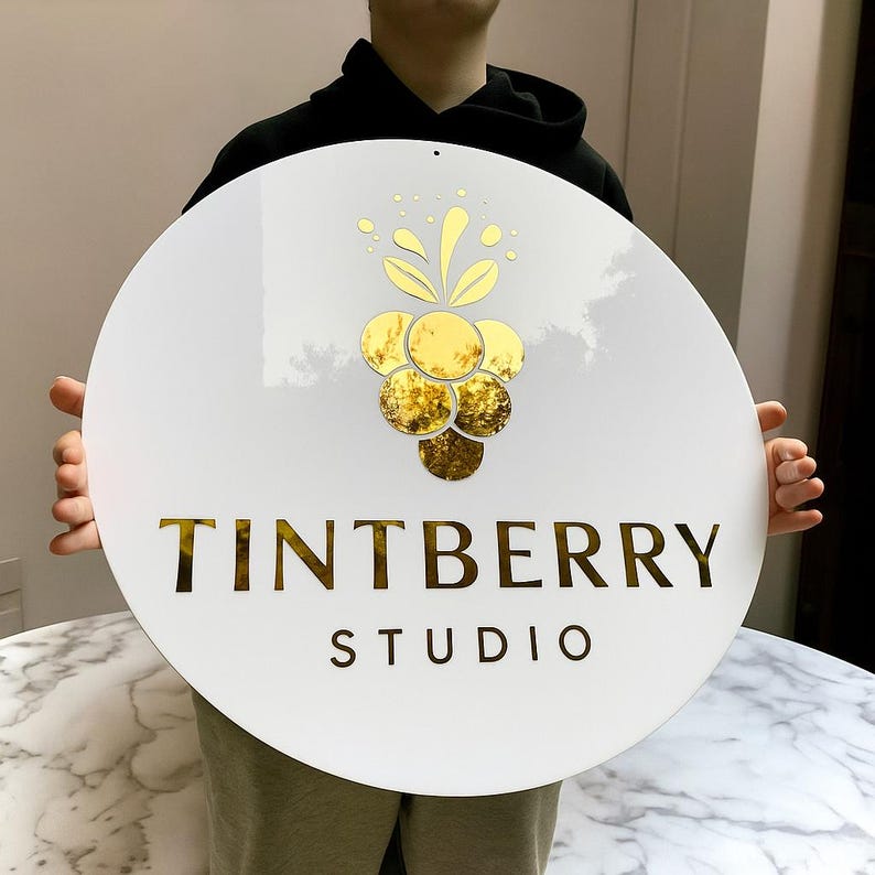 Może przedstawiać: Okrągły biały znak ze złotymi akcentami i napisem "TINTBERRY STUDIO". Znak przedstawia złoty wz&oacute;r grona winogron. Znak jest trzymany przez osobę.