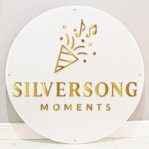 May include: A round white sign with gold lettering and accents. The text "SILVERSONG MOMENTS" is displayed in a bold, gold font. Above the text is a gold confetti popper and musical notes.