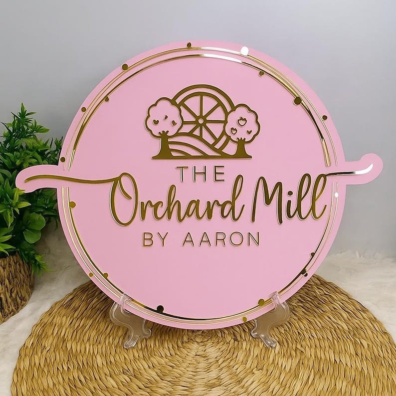 May include: A round pink sign with gold accents and the text "The Orchard Mill by Aaron." The sign features a gold illustration of a water wheel and trees. The sign is displayed on a clear stand.