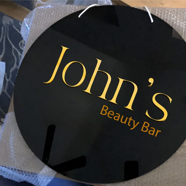 May include: Black circular sign with gold lettering that reads "John's Beauty Bar".