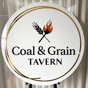 Może przedstawiać: Okrągły biały znak ze złotą obw&oacute;dką i czarnym napisem "Coal & Grain Tavern". Znak zawiera logo skrzyżowanych kłos&oacute;w pszenicy i płomienia w kolorze czarnym i pomarańczowym.