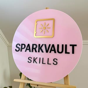 May include: A round, pink sign with the words "SPARKVAULT SKILLS" in black lettering. A gold-colored square with a sunburst design is at the top. The sign is displayed on a wooden easel.