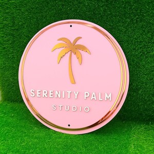Może przedstawiać: Okrągły, r&oacute;żowy znak z złotą obw&oacute;dką i złotym wzorem palmy. Tekst "SERENITY PALM STUDIO" jest w białych, dużych literach. Znak ma dwa małe otwory u g&oacute;ry do zawieszenia.
