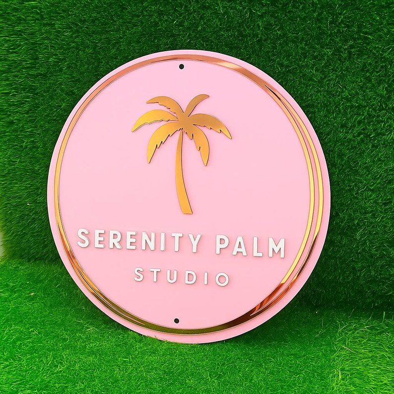 Może przedstawiać: Okrągły, r&oacute;żowy znak z złotą obw&oacute;dką i złotym wzorem palmy. Tekst "SERENITY PALM STUDIO" jest w białych, dużych literach. Znak ma dwa małe otwory u g&oacute;ry do zawieszenia.