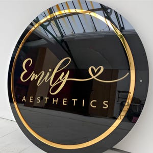 May include: A black circular sign with a gold border and gold lettering. The sign says "Emily Aesthetics" with a gold heart symbol.