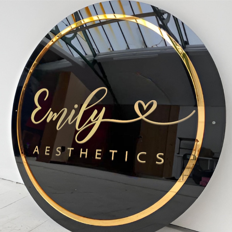 May include: A black circular sign with a gold border and gold lettering. The sign says "Emily Aesthetics" with a gold heart symbol.