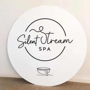 Pode incluir: Uma placa redonda branca com as palavras "Silent Stream SPA" em preto, com uma escrita elegante. O texto est&aacute; dentro de um c&iacute;rculo preto, com uma ilustra&ccedil;&atilde;o de uma tigela na parte inferior.