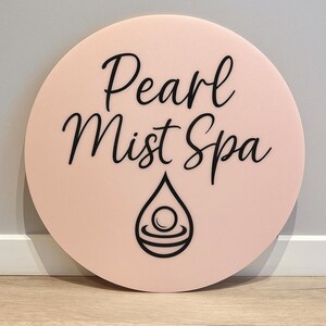 May include: A round, light pink sign with the words "Pearl Mist Spa" in black script. Below the text is a black teardrop design with a circular element inside. The sign is displayed against a neutral wall.