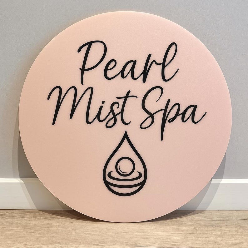 May include: A round, light pink sign with the words "Pearl Mist Spa" in black script. Below the text is a black teardrop design with a circular element inside. The sign is displayed against a neutral wall.
