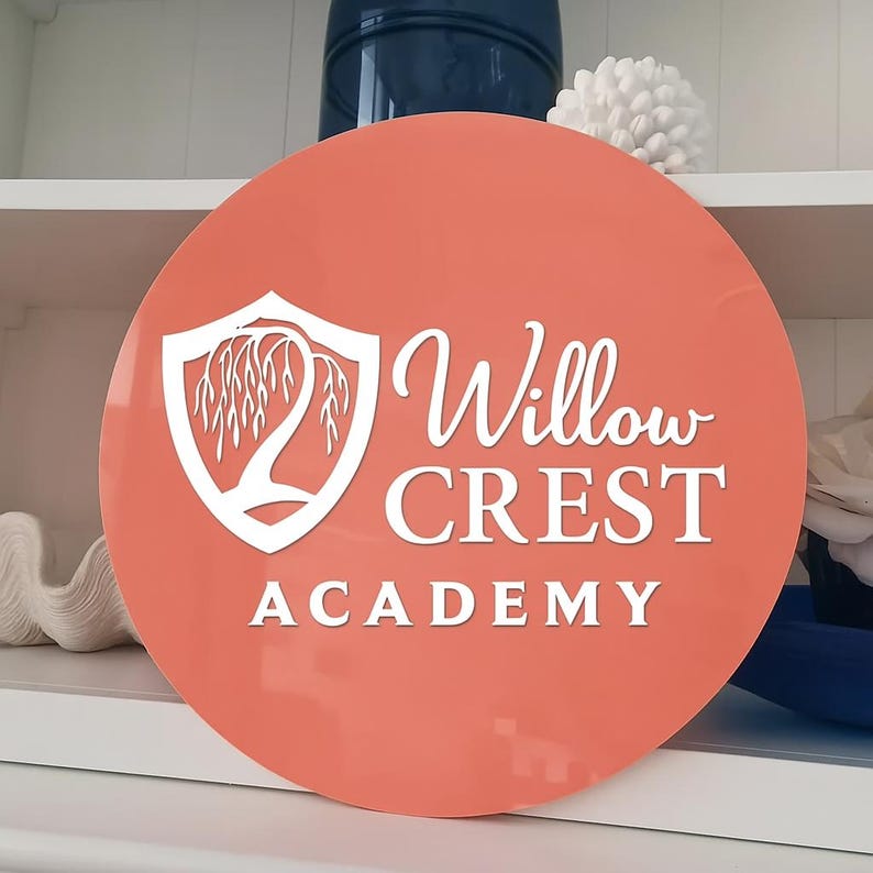 May include: A round, coral-colored sign with the text "Willow Crest Academy" in white script. The sign features a white shield with a stylized weeping willow tree design. The sign is displayed on a white shelf.