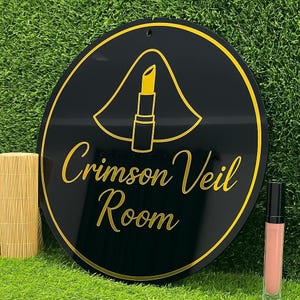 Pode incluir: Uma placa redonda preta e dourada com o texto "Crimson Veil Room" e um gr&aacute;fico de batom. Um tubo de brilho labial est&aacute; ao lado da placa. A placa est&aacute; em uma superf&iacute;cie verde.