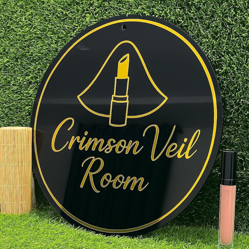 Pode incluir: Uma placa redonda preta e dourada com o texto "Crimson Veil Room" e um gr&aacute;fico de batom. Um tubo de brilho labial est&aacute; ao lado da placa. A placa est&aacute; em uma superf&iacute;cie verde.