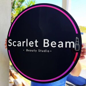 May include: A round black sign with a pink and magenta border. The sign reads "Scarlet Beam" in white text, with "Beauty Studio" below. A lipstick icon is on the right side of the sign.