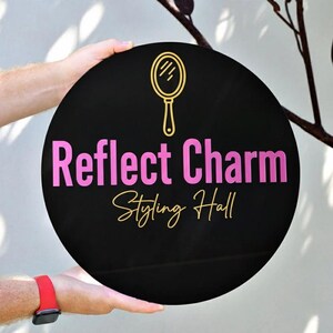 May include: A round black sign with the words "Reflect Charm Styling Hall" in pink and gold lettering. A gold mirror icon is above the text. The sign is held by a person.