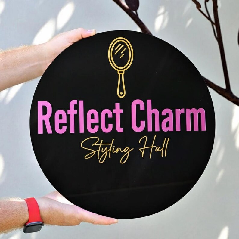May include: A round black sign with the words "Reflect Charm Styling Hall" in pink and gold lettering. A gold mirror icon is above the text. The sign is held by a person.