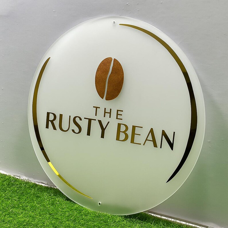 Może przedstawiać: Okrągły, biały znak z napisem "THE RUSTY BEAN" w złotych literach. Złoty graficzny motyw ziarna kawy znajduje się nad tekstem. Znak ma złotą obw&oacute;dkę i wisi na białej ścianie.