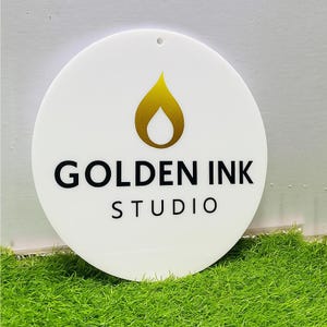 Puede incluir: Un letrero circular blanco que presenta un icono en forma de llama dorada sobre el texto negro "GOLDEN INK" y la palabra "STUDIO" centrada debajo. El letrero tiene un peque&ntilde;o orificio en la parte superior para colgar y una superficie lisa.