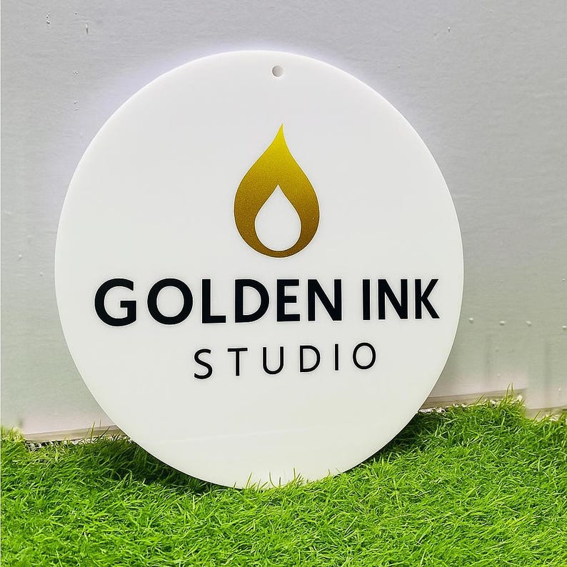 Puede incluir: Un letrero circular blanco que presenta un icono en forma de llama dorada sobre el texto negro "GOLDEN INK" y la palabra "STUDIO" centrada debajo. El letrero tiene un peque&ntilde;o orificio en la parte superior para colgar y una superficie lisa.