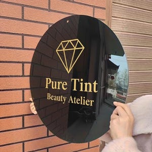 May include: A round, black sign with gold lettering and a diamond graphic. The sign reads "Pure Tint Beauty Atelier". The sign is held up against a brick wall.