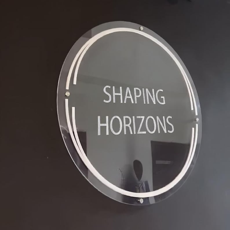 May include: A white circular sign with a double white ring border on a black background. The sign says "SHAPING HORIZONS" in white text.