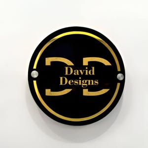 May include: A black circular sign with a gold border and the text "David Designs" in gold lettering. The letters "D" are stylized and connected.