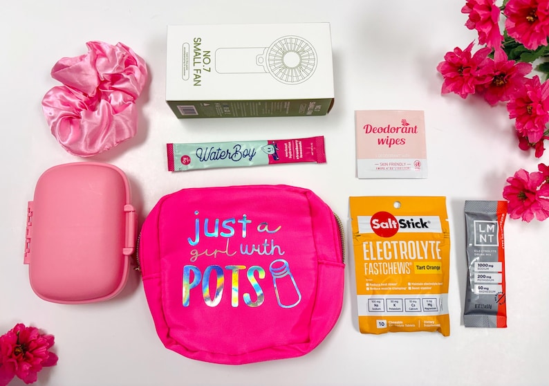 Puede incluir: Una colecci&oacute;n de accesorios y art&iacute;culos de salud rosas, que incluyen un ventilador peque&ntilde;o, una scrunchie, un estuche de viaje, una bolsa con el texto "just a girl with POTS", paquetes de electrolitos y toallitas desodorantes. Flores rosas en el fondo.