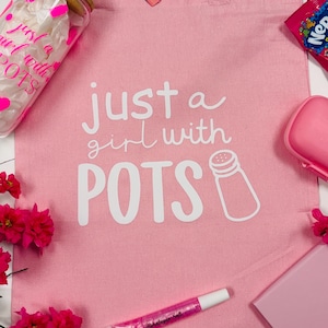 May include: A pink tote bag with the text "just a girl with POTS" in white. The bag is surrounded by pink accessories, including a lip gloss, a pink soap dish, and a jar with pink accents. A packet of electrolyte mix and a candy are also visible.