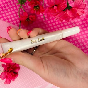 May include: A pearlescent lip gloss tube with the words "Sea Salt" in gold script. The tube has a white applicator tip and a gold charm. The background is pink with heart patterns and pink flowers.
