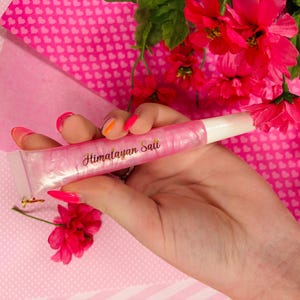 May include: A squeeze tube of lip gloss with a pearlescent pink finish and a white cap. The text "Himalayan Salt" is printed in a gold cursive font on the front of the tube. The tube is held against a background of bright pink heart patterns.