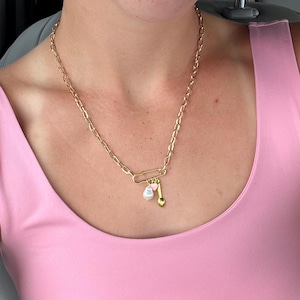 May include: Gold-toned chain necklace with a large paperclip-style clasp. The necklace features a pearl and a small gold-colored charm. The model is wearing a pink tank top.