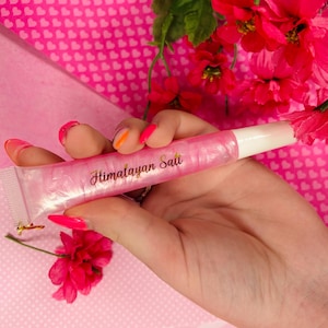 May include: A pink lip gloss tube with the text "Himalayan Salt" in gold lettering. The tube is held against a pink background with pink flowers.