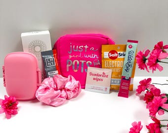 May include: A bright pink pouch with the text just a girl with POTS, a pink plastic case, a pink satin scrunchie, a white box, an LMNT electrolyte packet, a SaltStick electrolyte chew pouch, a deodorant wipe packet, and a Waterboy drink mix.