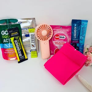 May include: A collection of items including a green GoSqueezy pouch, a yellow BodyArmor Flash I.V. stick, a silver cooling patch, a yellow Zero stick, a peach handheld fan, a pink SaltStick pouch, a blue Liquid I.V. stick, and a bright pink fabric.