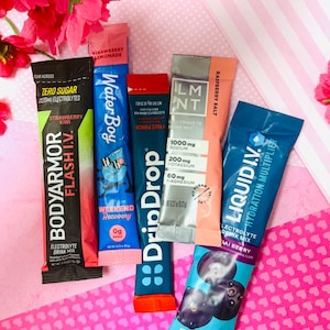 May include: Assortment of electrolyte drink mix packets in various colors, including pink, blue, and orange. Brands include BodyArmor, Water Boy, DripDrop, LMNT, and Liquid I.V. Flavors include strawberry kiwi, fruit punch, and raspberry salt. The packets are arranged on a pink patterned surface.