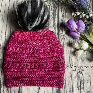 May include: A handmade, magenta-colored crochet beanie with a black and white faux fur pom-pom. The hat features a textured stitch pattern and is displayed on a wooden surface. The beanie is a winter accessory.