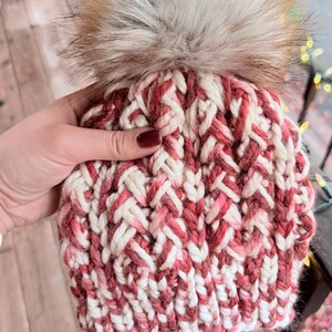 May include: A hand-knitted winter hat with a cream, red, and pink woven pattern. The hat features a large, fluffy, faux fur pom-pom on top. The hat is made of thick yarn, perfect for cold weather.