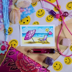 May include: A collection of decorative items including a small watercolor painting of a beach scene with a pink umbrella, a pink beaded necklace, a pink and purple pouch, and a clear tube with a dark pink interior. The background includes a fabric with yellow smiley faces.